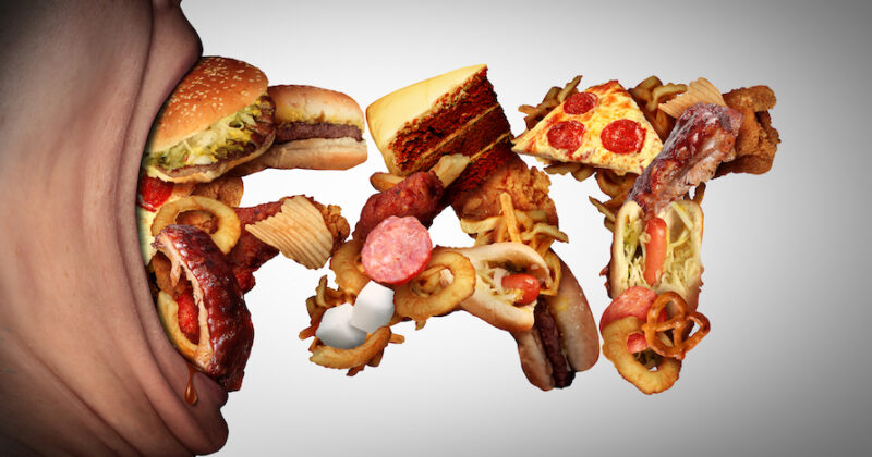 Eating fat food concept as an open mouth biting into unhealthy snacks shaped as text as a bad nutrition and obesity symbol for craving high calorie fatty meals.